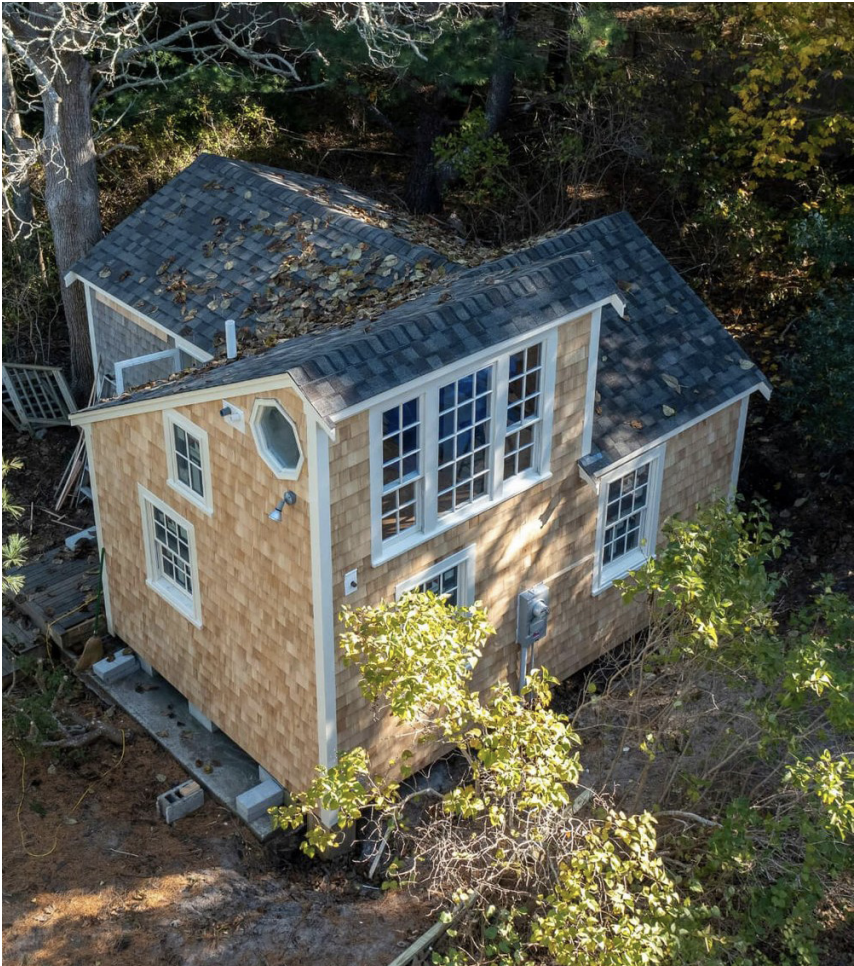 A birds-eye photograph of a 1.5-story accessory dwelling unit. The home has wooden shingle walls and slate shingle roof with traditional windows, including a large set of windows in the loft area. The unit sits on a raised foundation over a slab. The home is surrounded by trees and brush.